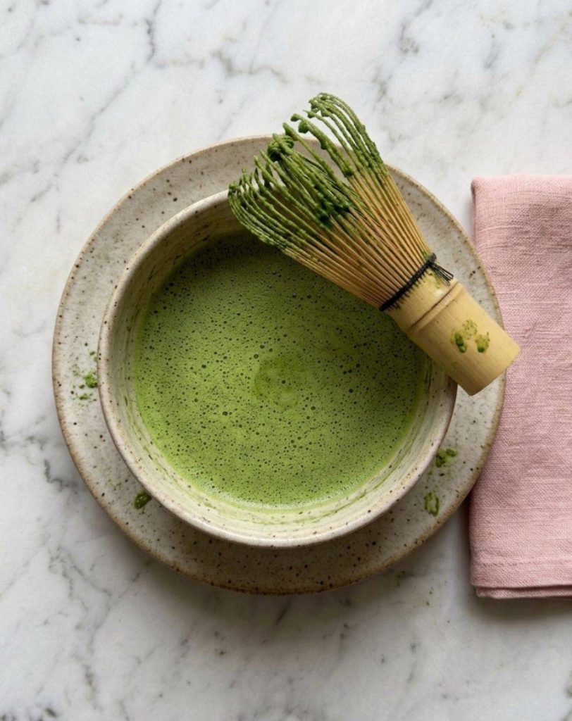 Bol de matcha fouetté avec une mousse fine couleur jade, un chasen en bambou maculé de matcha posé sur le bord et un chakin plié sur le plan de travail.